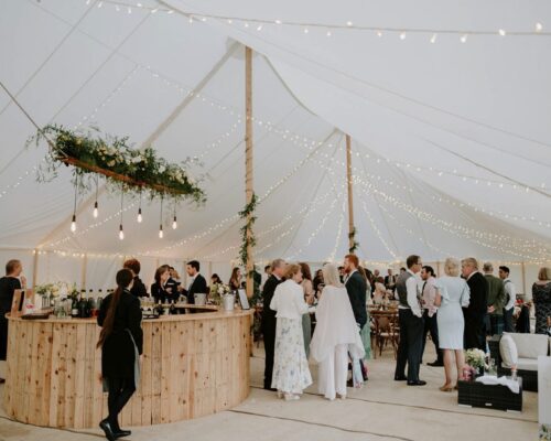 Elegant traditional pole marquee with festoon lighting, a rustic wooden bar, and guests enjoying a sophisticated wedding celebration.