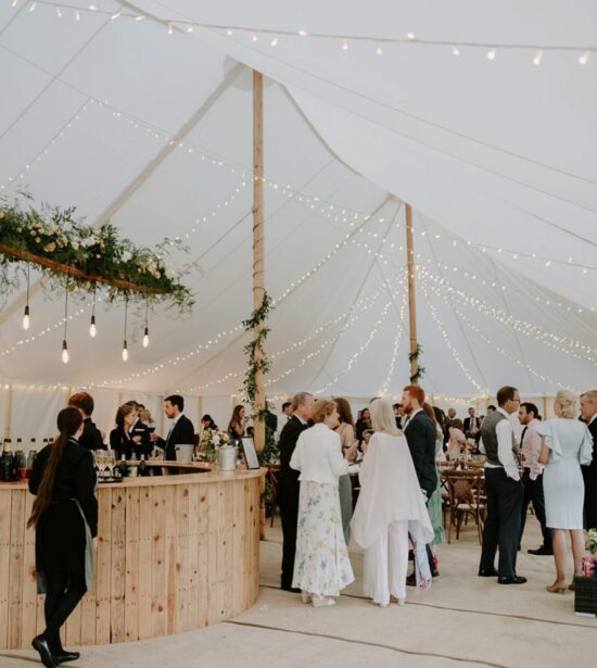 Elegant traditional pole marquee with festoon lighting, a rustic wooden bar, and guests enjoying a sophisticated wedding celebration.